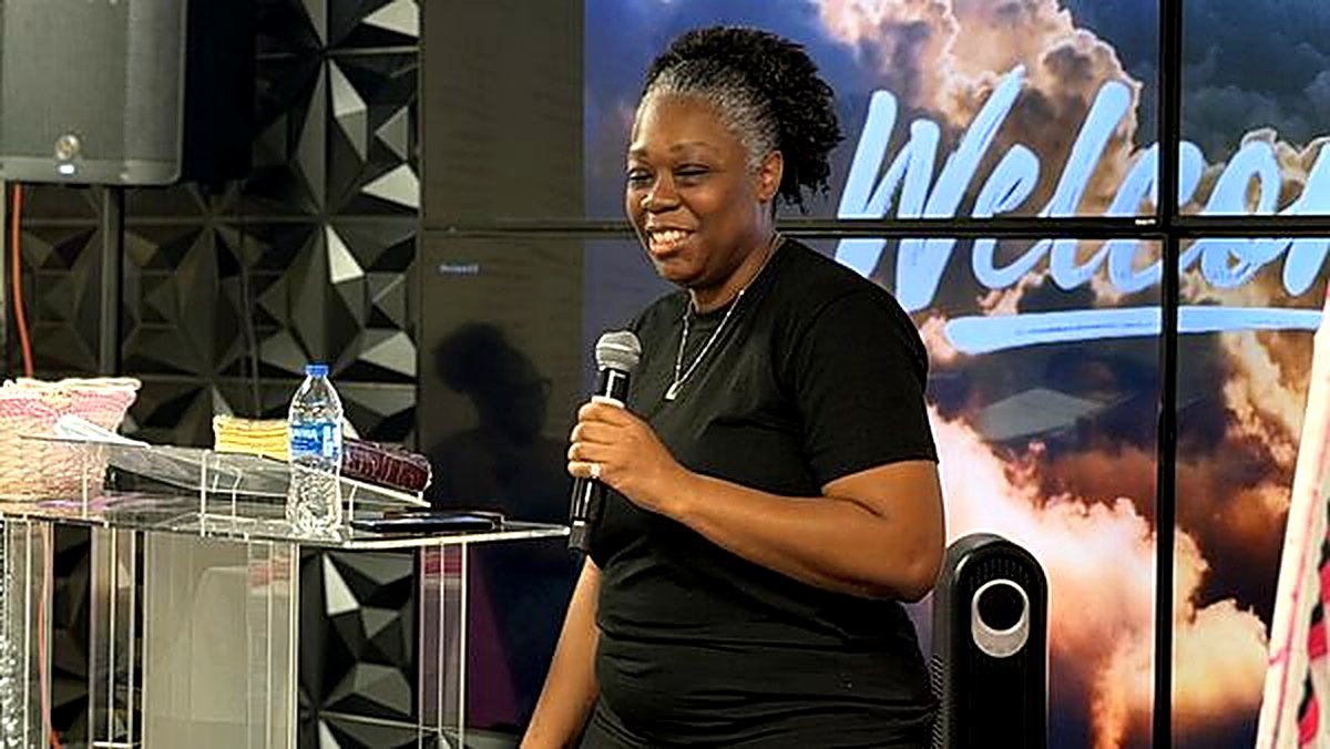 Pastor Helen ministering with a microphone in front of the Welcome screen